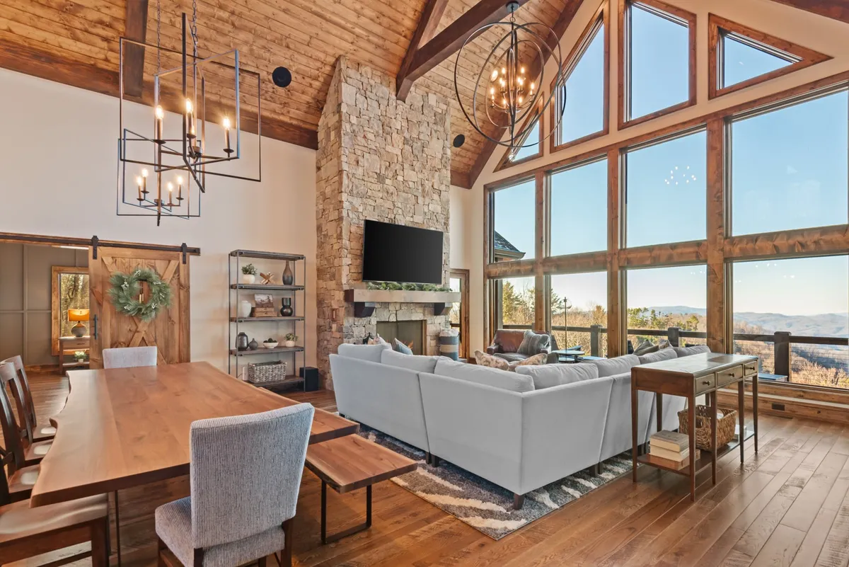 An inviting great room with a large stone fireplace as the focal point, expansive panoramic windows, a live-edge dining table, and a comfortable sectional sofa. The space is bathed in natural light and offers beautiful mountain views, perfect for family gatherings, casual dining, and watching movies on an impressive TV.
