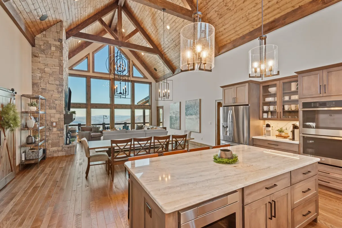This image features a spacious, open-plan kitchen with a prominent granite island, stainless steel appliances, and modern light fixtures. Expansive windows flood the space with natural light and offer scenic views. The adjacent dining area showcases a wooden table ideal for group gatherings, while exposed wooden beams add mountain charm to the ambiance.
