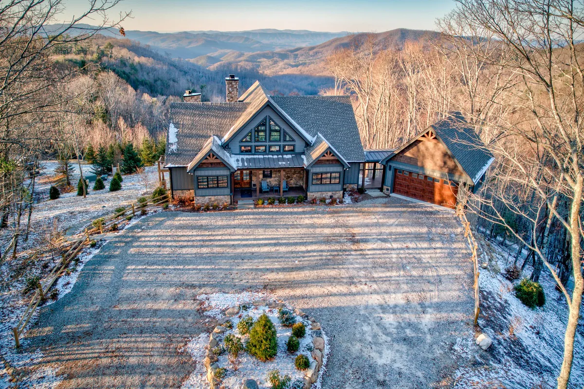 The image showcases a striking, modern mountain home set against a breathtaking backdrop of rolling North Carolina hills. The expansive driveway draws the eye toward the home, which features inviting porches perfect for enjoying sunset views. Lush greenery and the promise of nearby nature trails surround the property, evoking a sense of peace and adventure.