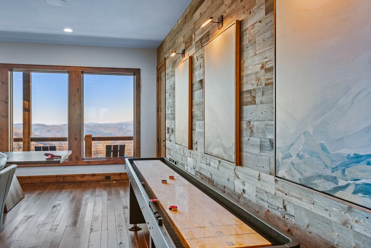 A bright, fun game room with a shuffleboard table running along a wooden wall. Large sliding windows offer scenic mountain views, and the space feels open and energetic, inviting guests to play and socialize.
