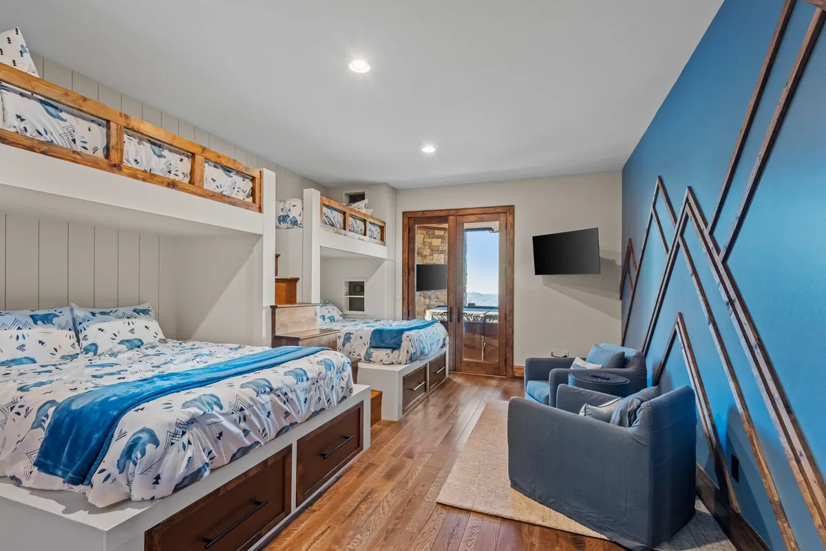 A playful and comfortable bunkroom with a combination of bunk beds and queen beds, a blue accent wall, and wooden furniture. The room is arranged for group fun and family bonding, offering cozy spaces for sleeping, watching TV, playing games, and sharing stories. Natural light fills the space, creating a cheerful, welcoming environment.