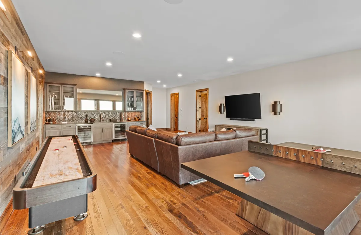 A vibrant interior scene featuring a spacious entertainment lounge. The room is centered around a large leather sectional sofa facing a big-screen TV, with a bar area and a ping pong table visible. The atmosphere is casual and inviting, perfect for group gatherings and relaxing game nights.