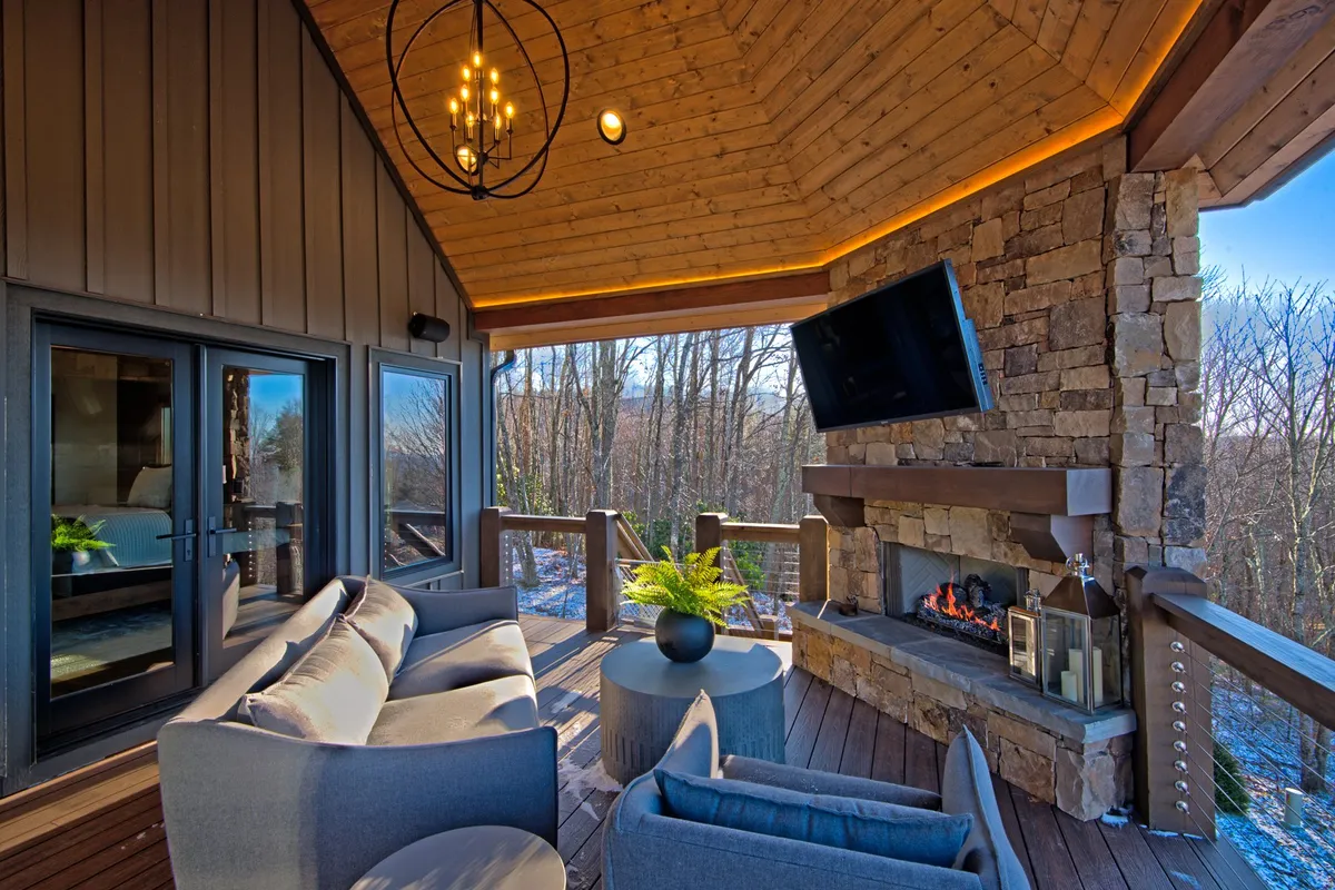 A spacious and stylish covered porch features a comfortable seating area, a stone fireplace, and large windows with views into a tranquil forest. The setting is perfect for shared laughter, group gatherings, and enjoying peaceful evenings outdoors.