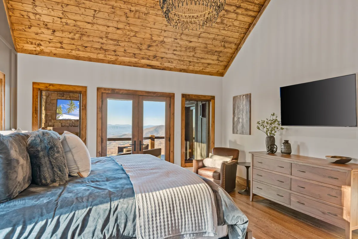 This image features a king-sized bed with crisp white bedding, positioned beside expansive windows that frame a stunning mountain view. A comfortable armchair rests in one corner, and the room is decorated with tasteful wall art and a wooden dresser, all illuminated by soft, natural light. The atmosphere is serene and cozy, inviting relaxation and private comfort.