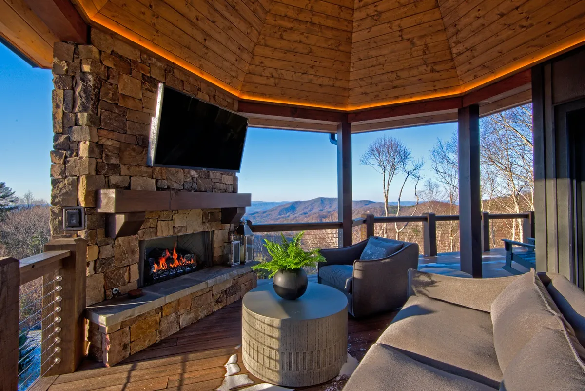 A beautifully designed outdoor deck featuring a stone fireplace, a large TV mounted above, comfortable lounge seating, a coffee table with a plant, and sweeping mountain views beyond the railing. The scene is set for relaxation, socializing, and enjoying both technology and nature in equal measure.