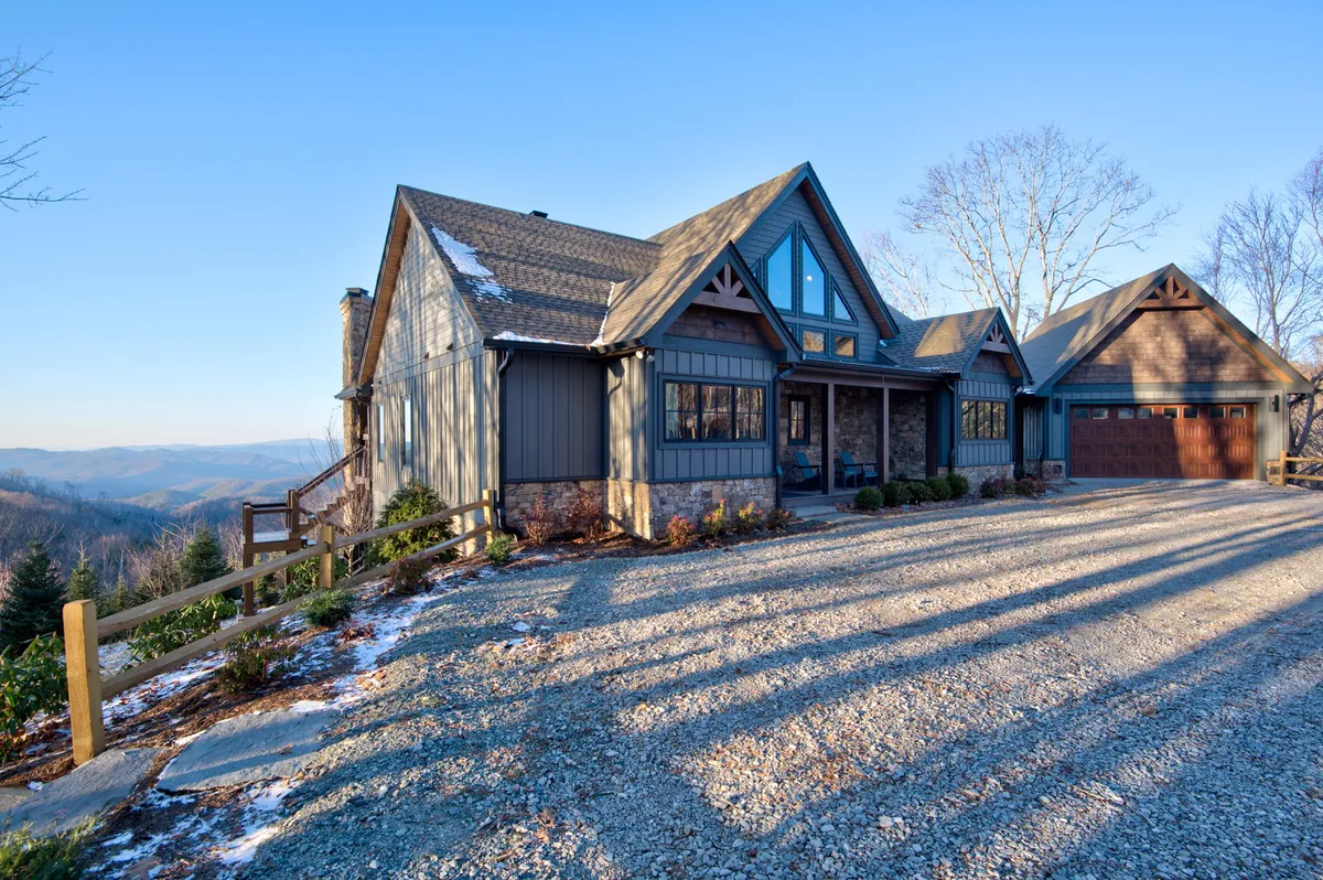 The photo features the main entrance to a grand house with a distinctive stone facade and unique roof, framed by mountain vistas. The scene conveys a sense of arrival and the promise of both communal and private family experiences in a mountain retreat.
