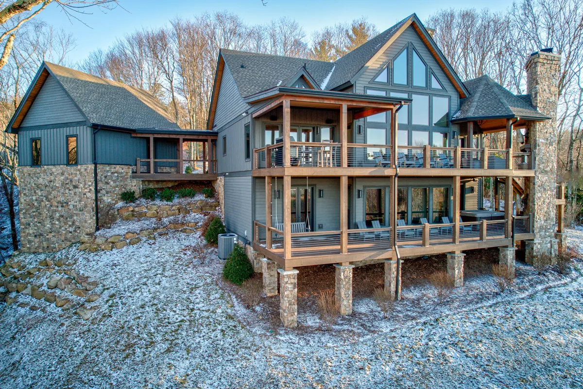 This exterior shot highlights large glass windows, multiple wooden decks, and stone accents, all set against a backdrop of mature trees. The image communicates opportunities for outdoor gatherings, relaxation on the deck, and immersing in nature, reflecting both summer and transitional seasons at the property.